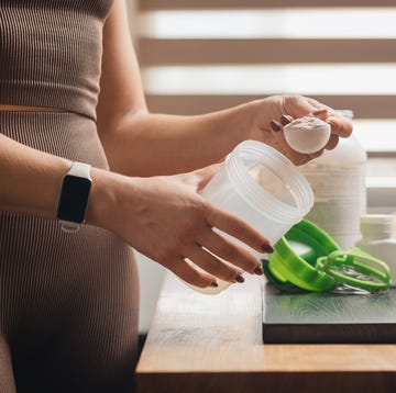 athletic woman in sportswear with measuring spoon in her hand puts portion of whey protein powder into a shaker on wooden table with amino acid white capsules, bananas and apple, making protein drink