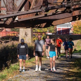 group of people walking and running along a path under a rusty bridge